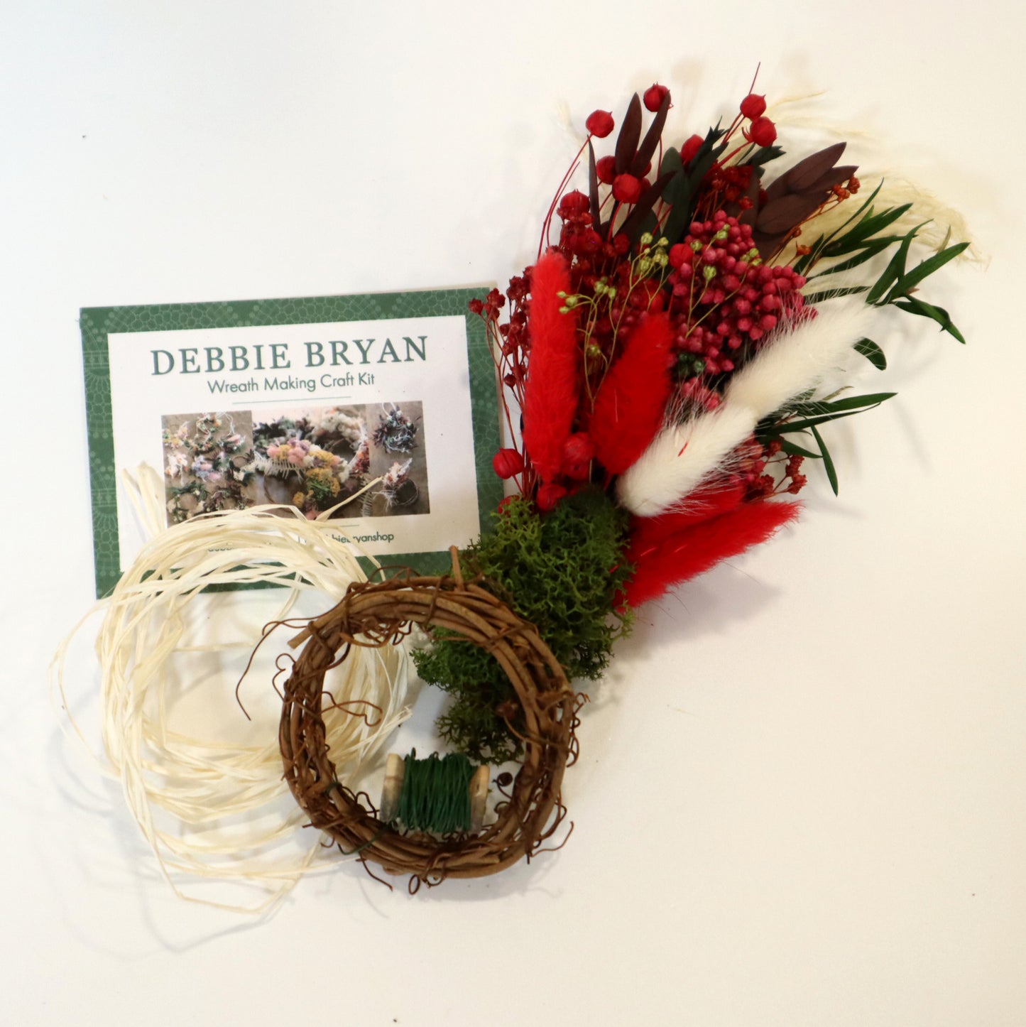 Wreath making craft kit with materials and instructions on a white background, including a small dried flower bunch, preserved green moss, a small vine wreath, floristry wire and raffia