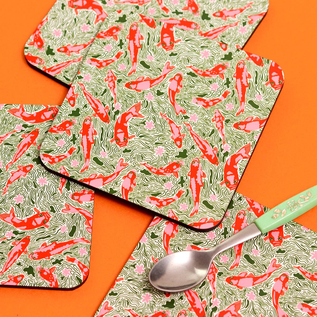 Coasters featuring a pink and red fish pattern on a green water background. Sat on an orange background