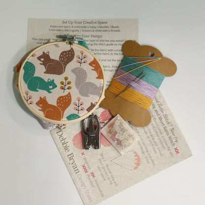 Contents of a craft kit including squirrel themed embroidery hoop, a how to guide, embroidery threads in yellow, blue and purple, folding scissors and beads