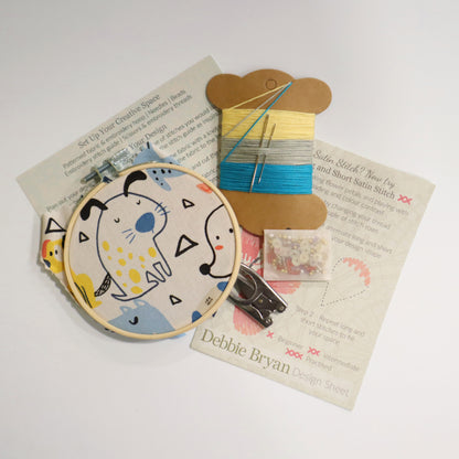 Contents of a craft kit including dog themed embroidery hoop, a how to guide, embroidery threads in yellow and blues, folding scissors and beads
