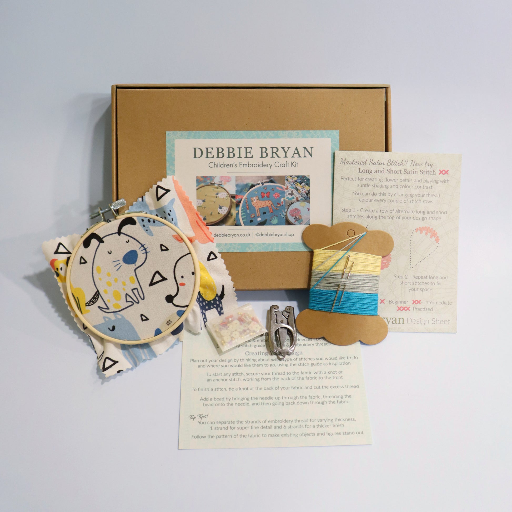 Dog themed children's embroidery craft kit with a cardboard box, hoop, patterned fabric, and instructions on a light grey background.