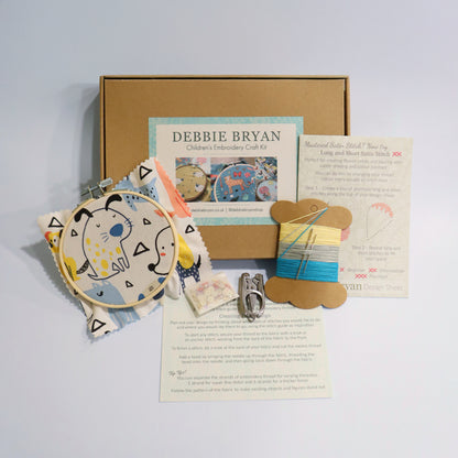 Dog themed children's embroidery craft kit with a cardboard box, hoop, patterned fabric, and instructions on a light grey background.