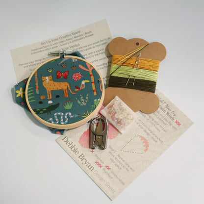 Contents of a craft kit including jungle themed embroidery hoop, a how to guide, embroidery threads in yellow and greens, folding scissors and beads