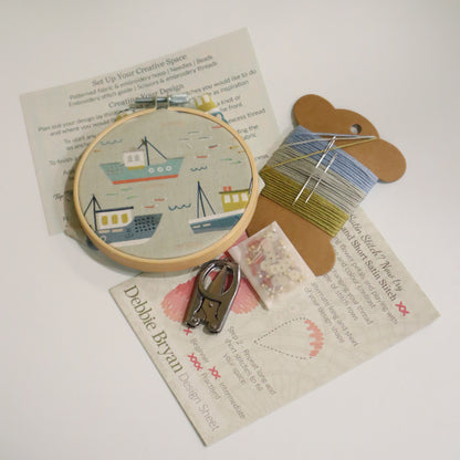 Contents of a craft kit including boat themed embroidery hoop, a how to guide, embroidery threads in blues and green, folding scissors and beads