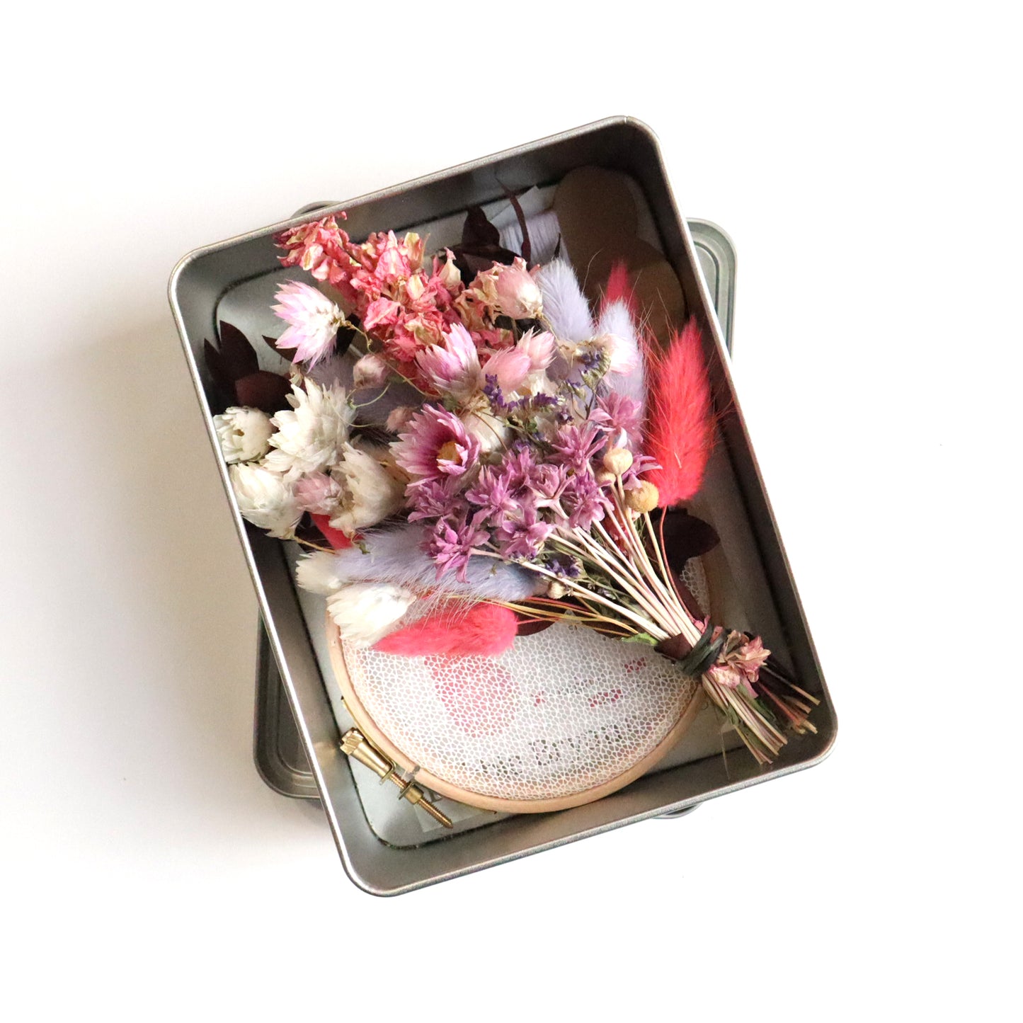 Metal tin with a pink dried flower bunch and Nottingham Lace tulle sat in an embroidery hoop on a white background