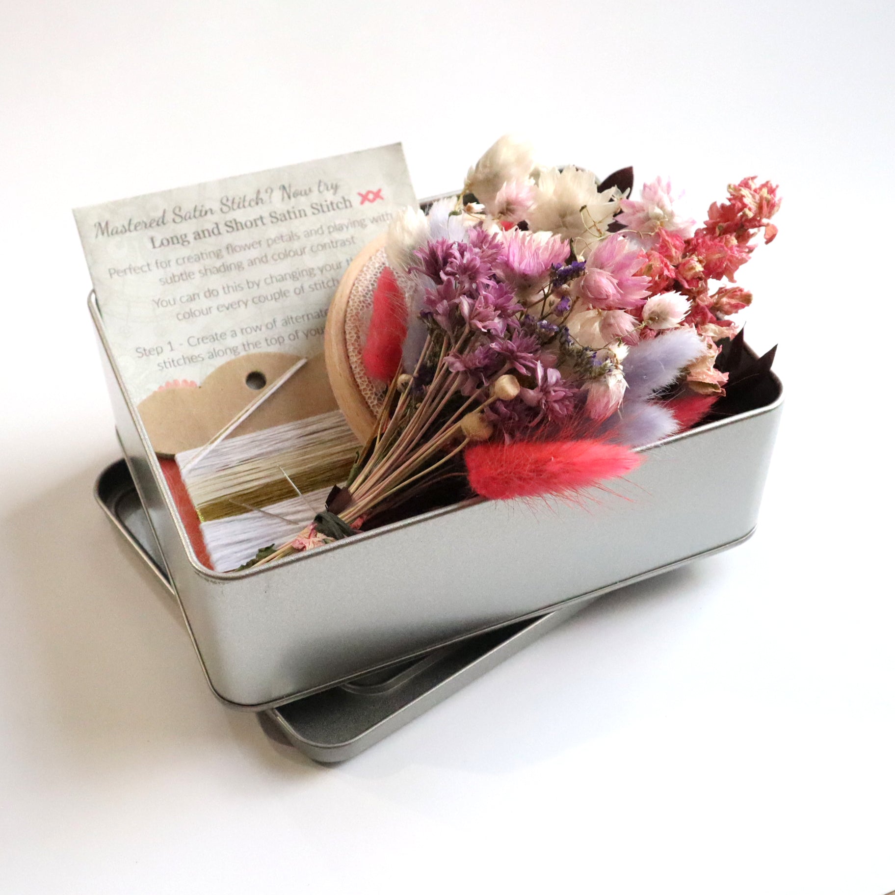 Metal tin which contains a pink dried flower bunch, embroidery threads and a how-to stitch guide