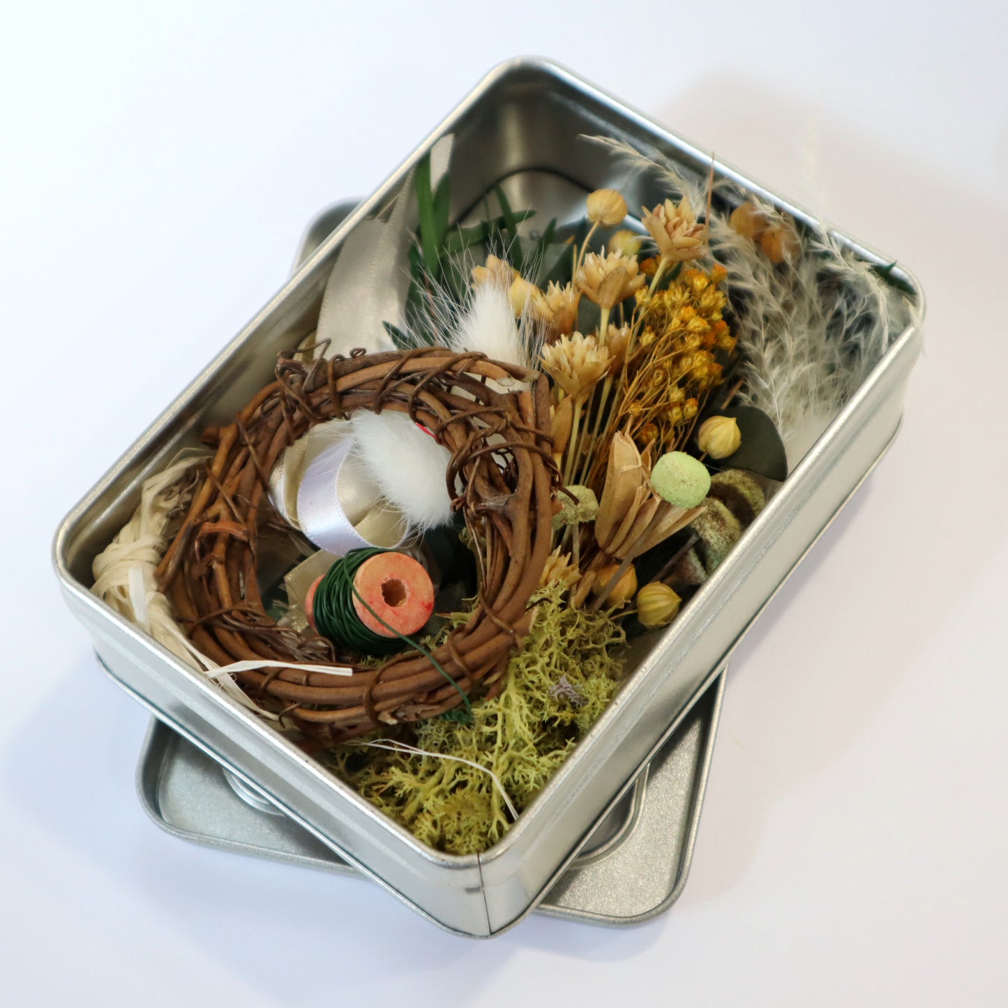 Metal tin with a small vine wreath, green wire reel and a small bunch of dried flowers on a white background