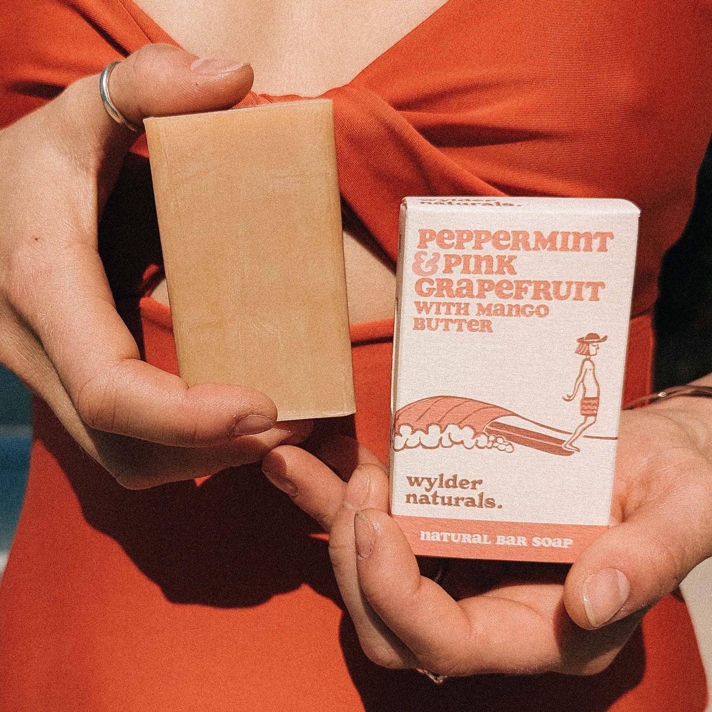 A bar of soap labeled 'Peppermint & Pink Grapefruit with Mango Butter Natural Soap' being held by a person with a beach and ocean in the background.