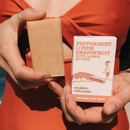 A bar of soap labeled 'Peppermint & Pink Grapefruit with Mango Butter Natural Soap' being held by a person with a beach and ocean in the background.