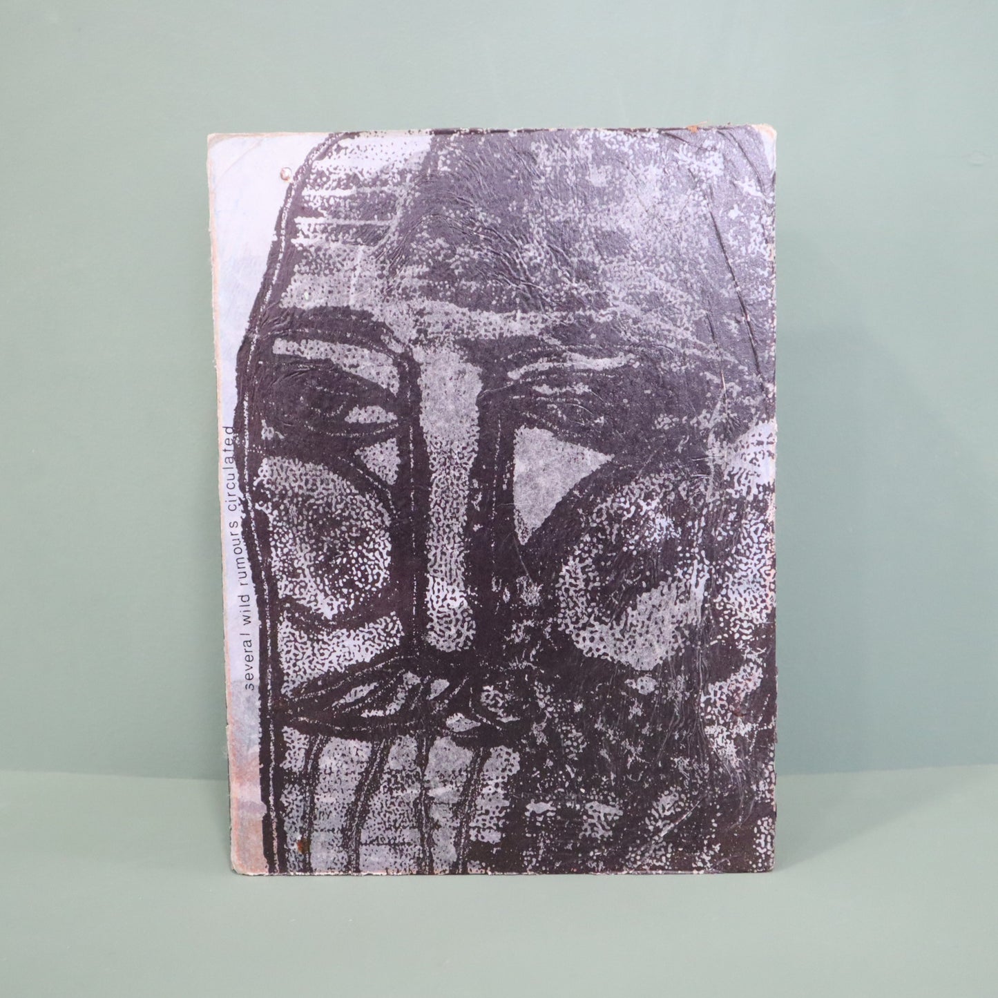 Mixed media artwork with a textured surface featuring a stylized face design on a light green background