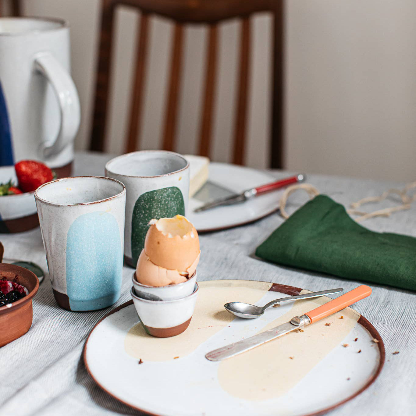 Dining table with ceramic cups, an eggshell, a plate with a knife and spoon, and a mug.