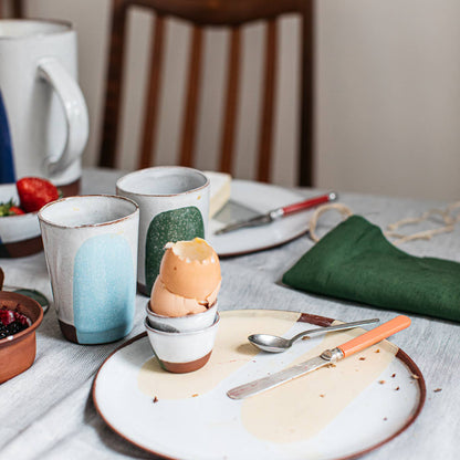 Dining table with ceramic cups, an eggshell, a plate with a knife and spoon, and a mug.