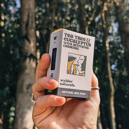 A hand holding a bar of soap with packaging that includes an illustration of a plant and text detailing the product type and brand name.