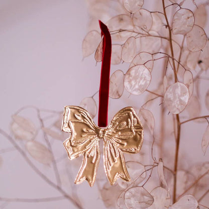 A tin embossing bow ornament in the shape of a bow with a red ribbon, displayed on a branch with a blurred background.