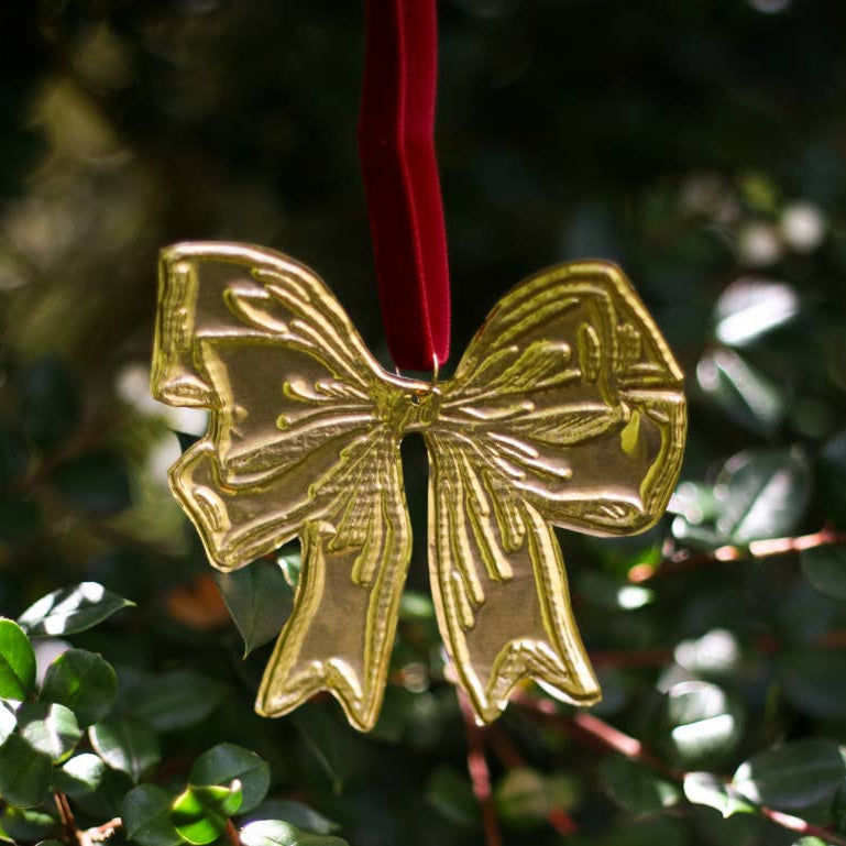 A tin embossing bow ornament in the shape of a bow with a red ribbon, displayed on a branch with a blurred green background.