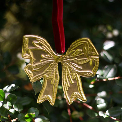 A tin embossing bow ornament in the shape of a bow with a red ribbon, displayed on a branch with a blurred green background.