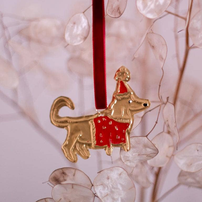 A tin embossing dog in jumper ornament with a red ribbon, placed on a branch with leaf-like decorations in the background.