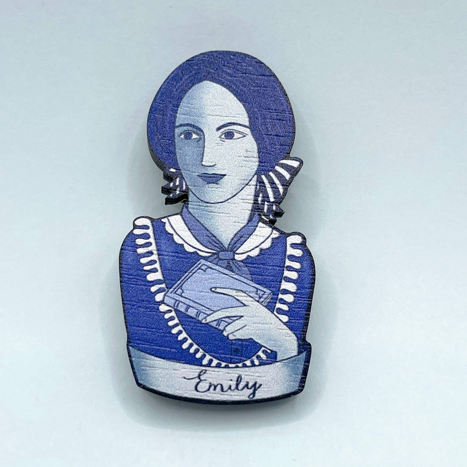 Brooch with a blue and white design of Emily Bronte holding a book, on a light gray background.