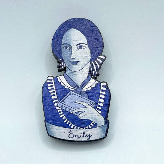 Brooch with a blue and white design of Emily Bronte holding a book, on a light gray background.