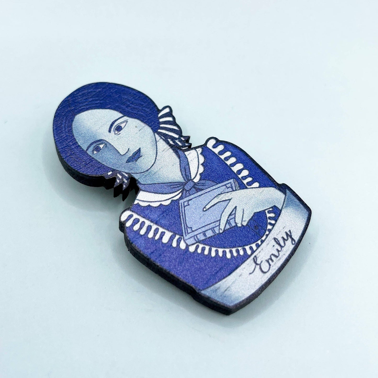 Brooch with a blue and white design of Emily Bronte holding a book, on a light grey background.