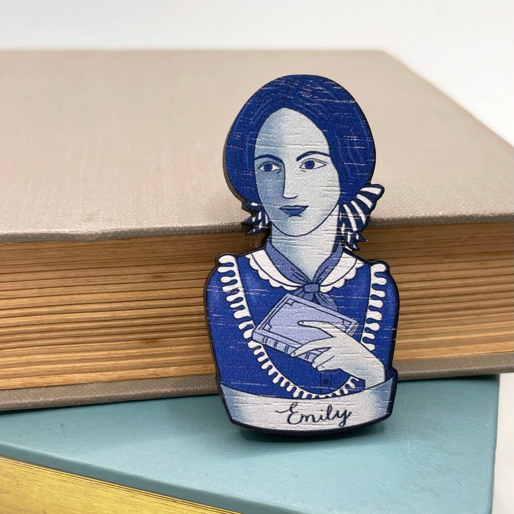 Brooch with a blue and white design of Emily Bronte holding a book, sitting on a stack of books