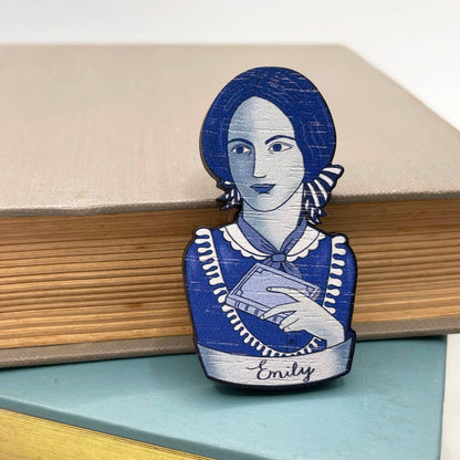 Brooch with a blue and white design of Emily Bronte holding a book, sitting on a stack of books