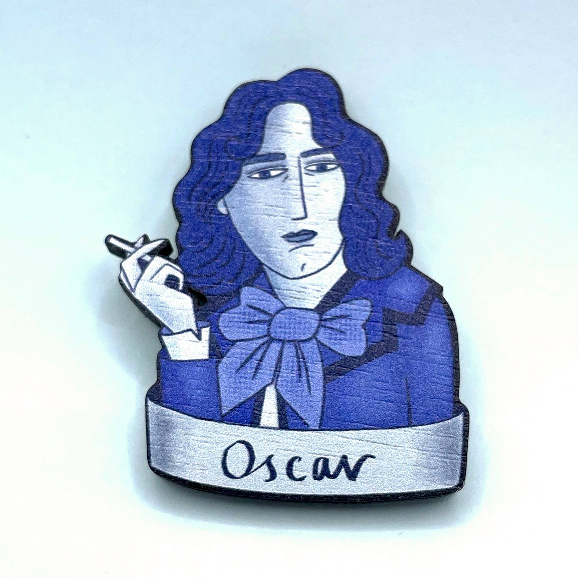 A wooden brooch depicting Oscar Wilde with a blue bow tie, smoking a cigarette.