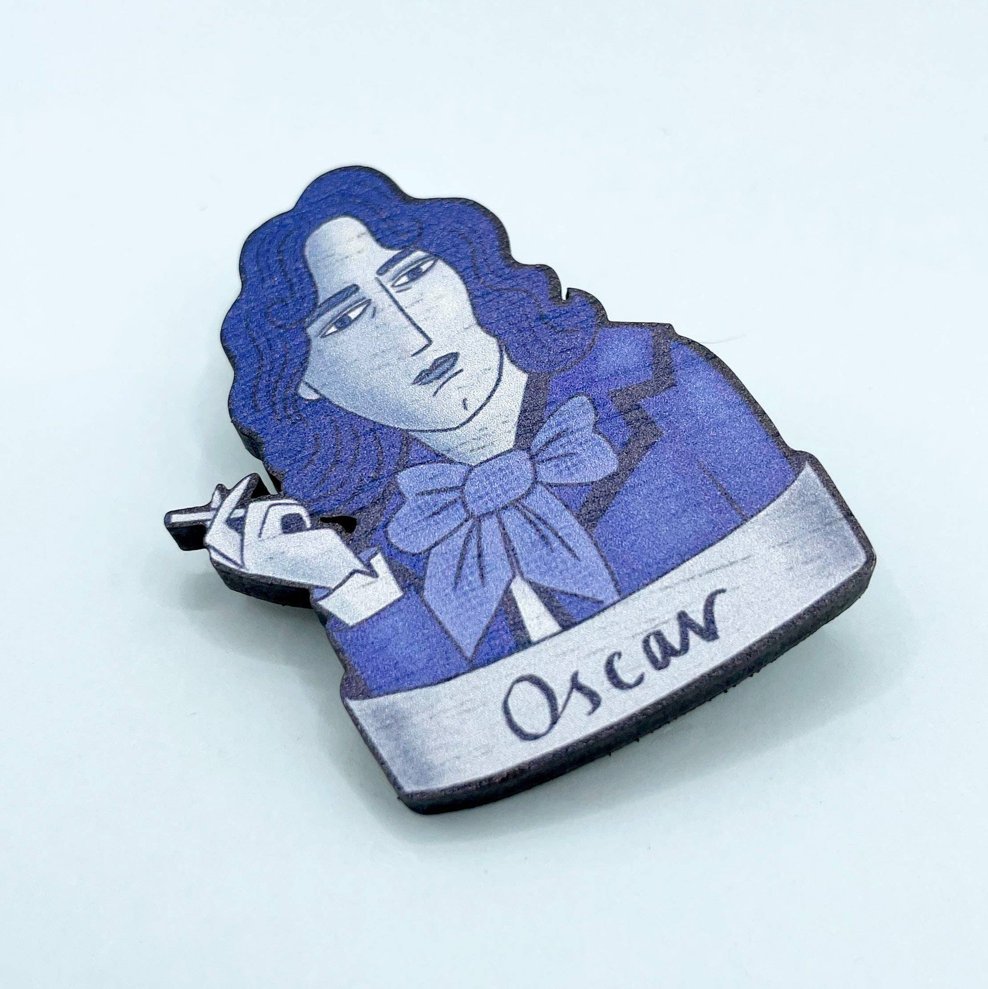 A wooden brooch depicting Oscar Wilde with a blue bow tie, smoking a cigarette.