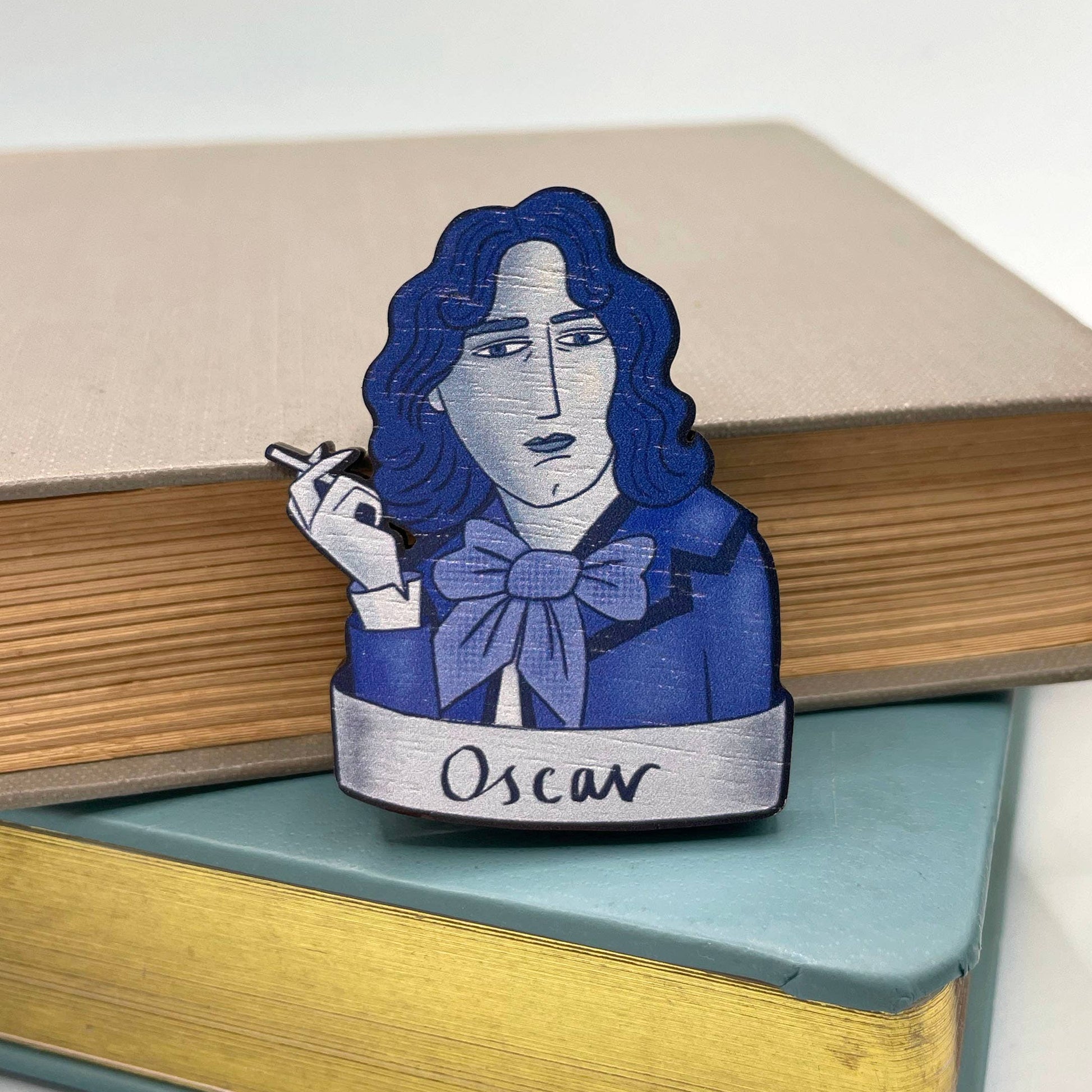 A wooden brooch depicting Oscar Wilde with a blue bow tie, smoking a cigarette sitting on a stack of books
