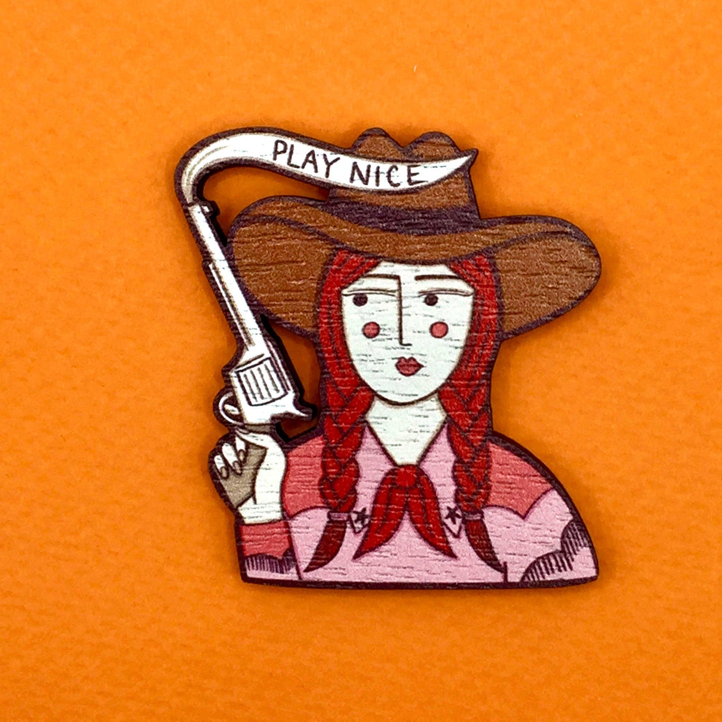 A brooch depicting a cartoon cowgirl with a gun and the phrase 'PLAY NICE' on a brown background.