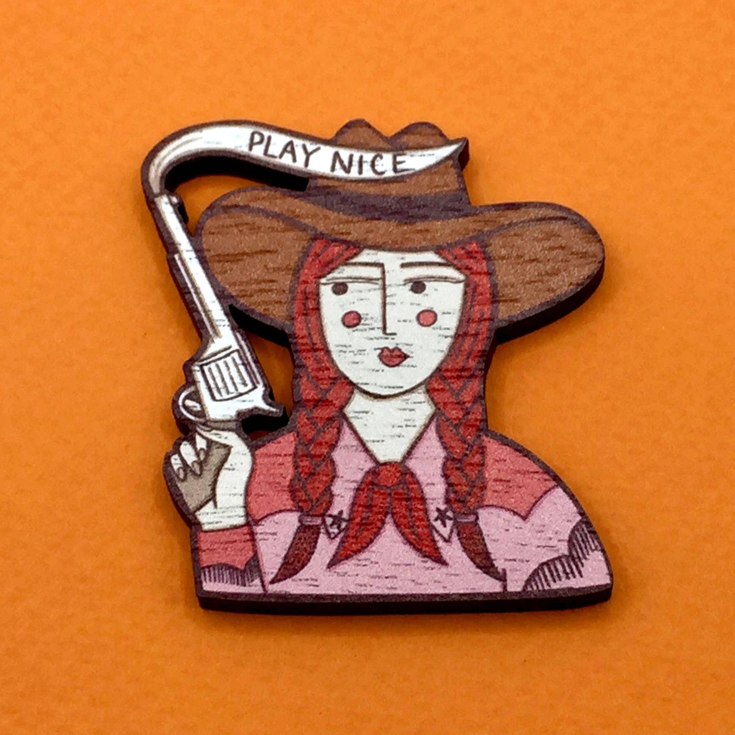 Wooden badge with a design of a woman in a cowboy hat holding a gun, on an orange background.