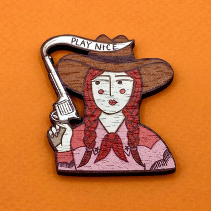 Wooden badge with a design of a woman in a cowboy hat holding a gun, on an orange background.