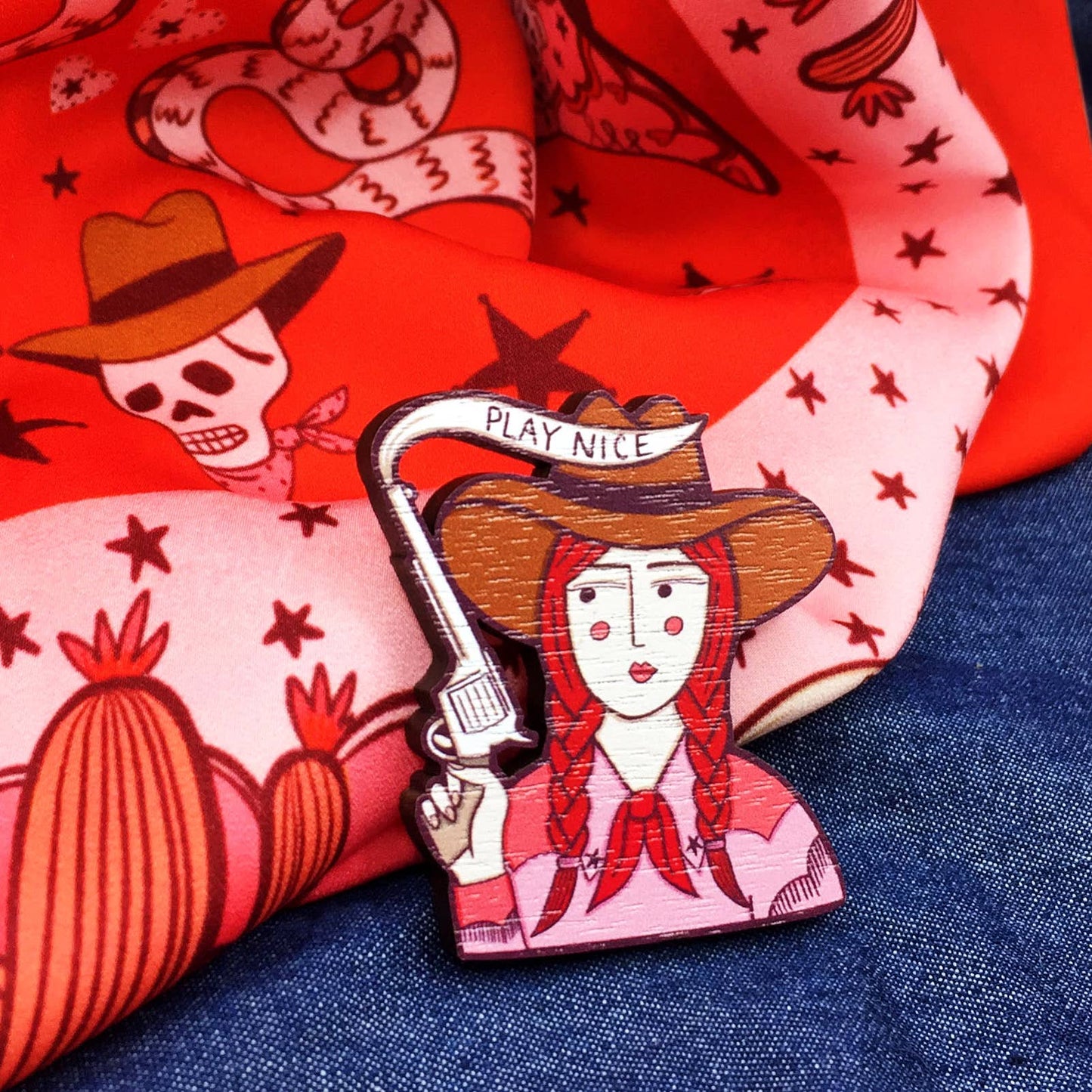A brooch depicting a cartoon cowgirl with a gun and the phrase 'PLAY NICE' on denim and pink patterned fabric background