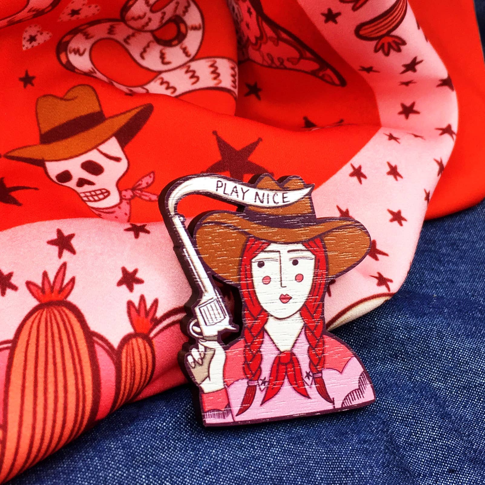 A brooch depicting a cartoon cowgirl with a gun and the phrase 'PLAY NICE' on denim and pink patterned fabric background