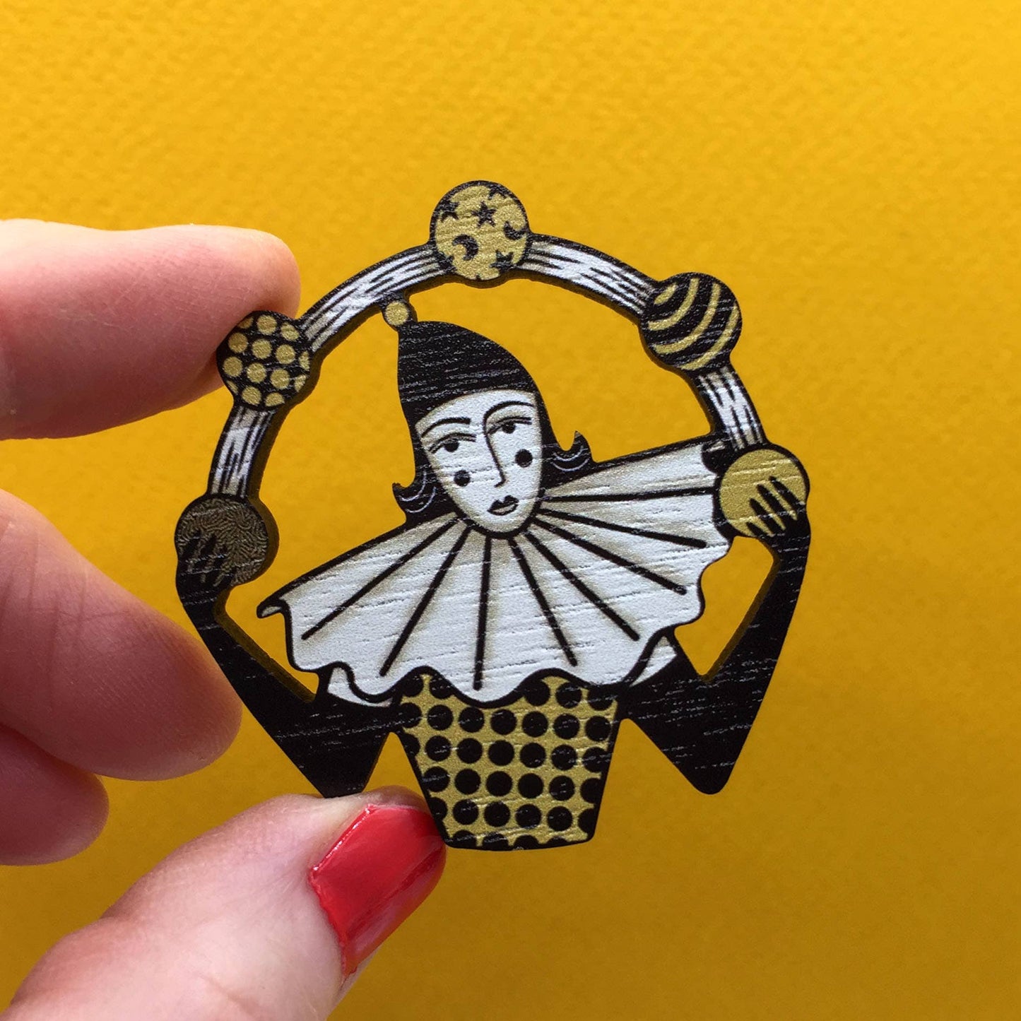 A hand holding a wooden pin brooch depicting a vintage style circus juggler, with a sombre look, laser-cut from sustainable walnut wood.