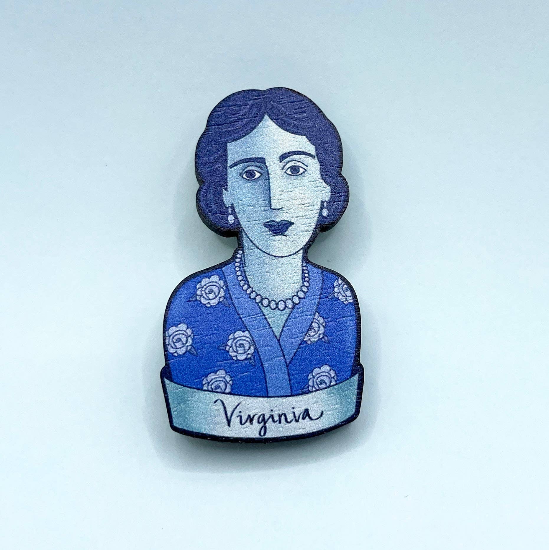A wooden brooch depicting Virginia Woolf with a blue dress and rose motifs, secured by a metal bar pin.