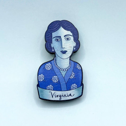 A wooden brooch depicting Virginia Woolf with a blue dress and rose motifs, secured by a metal bar pin.