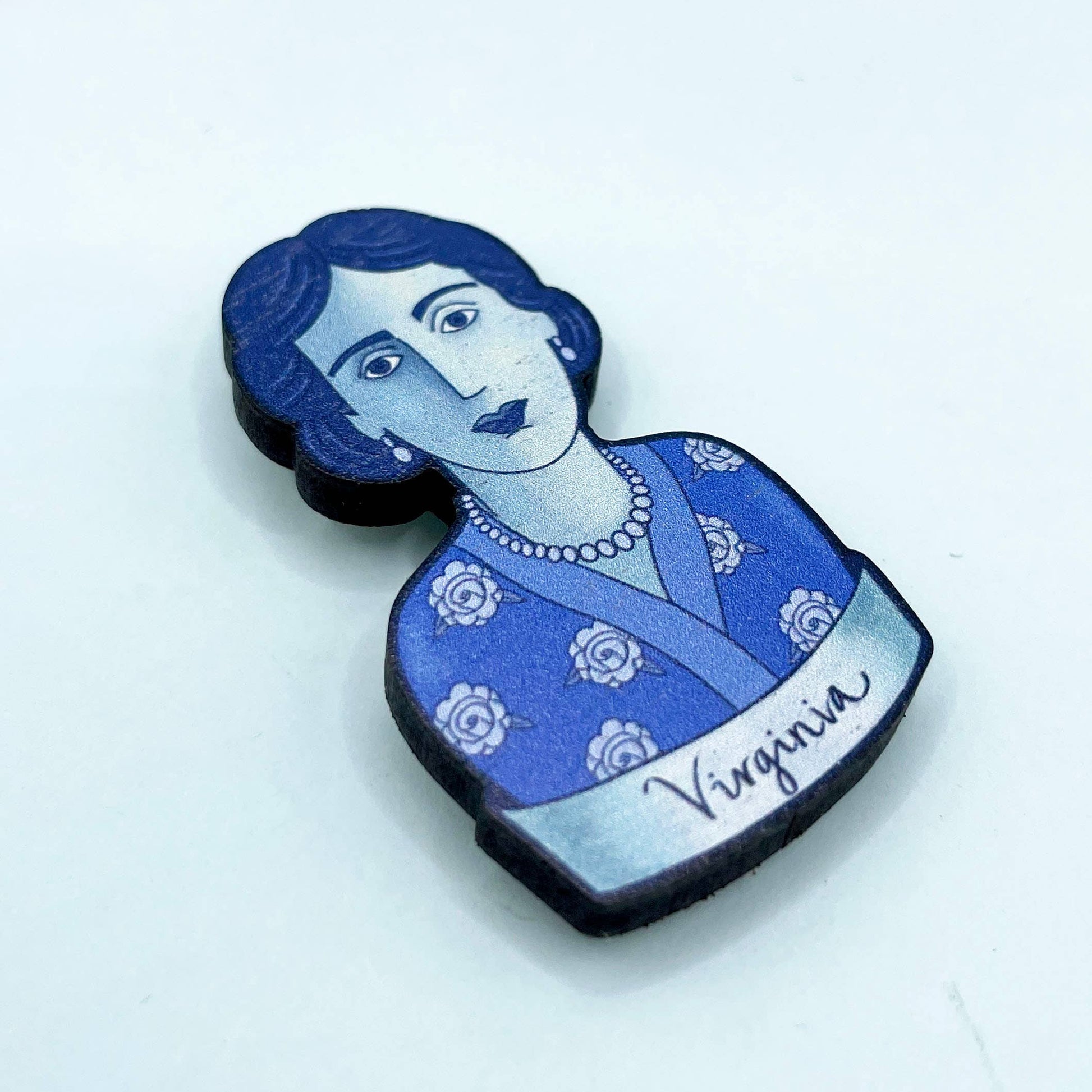 A wooden brooch depicting Virginia Woolf with a blue dress and rose motifs, secured by a metal bar pin.