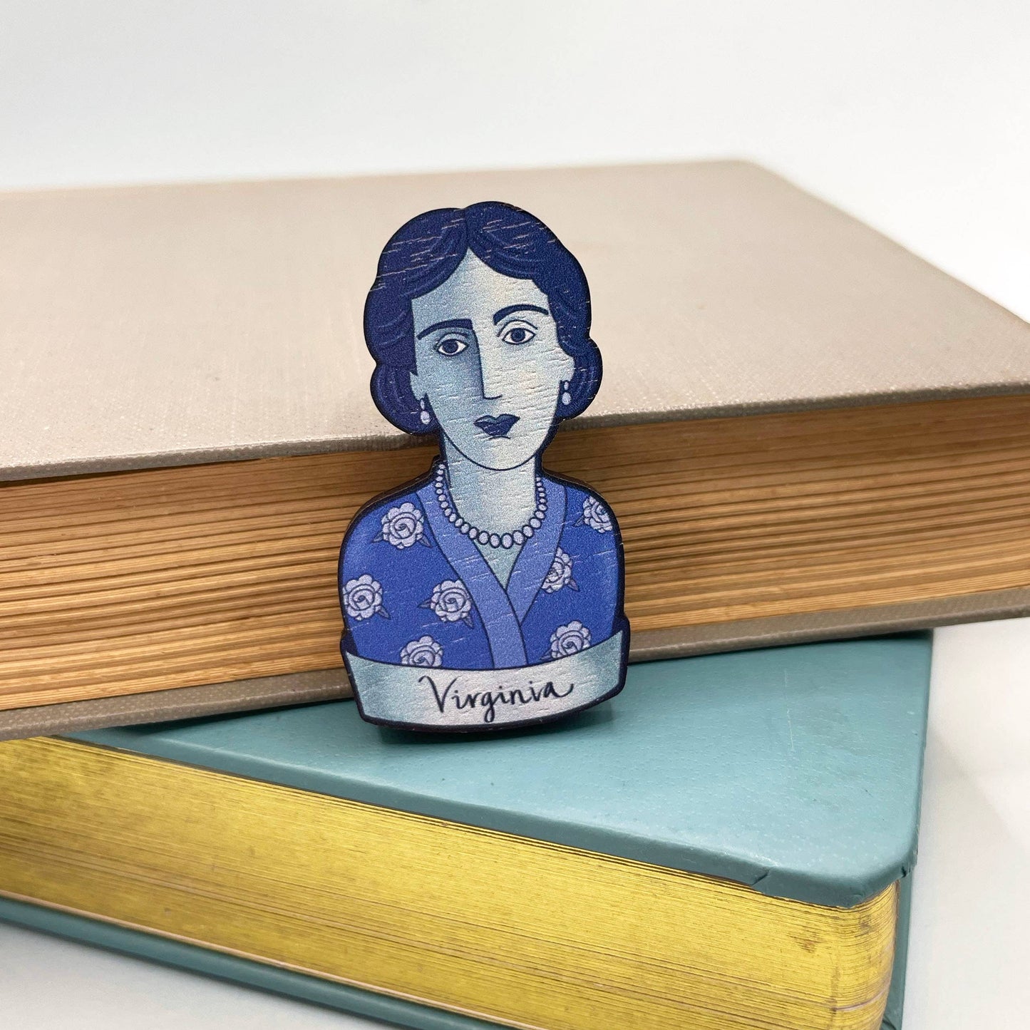 A wooden brooch depicting Virginia Woolf with a blue dress and rose motifs, secured by a metal bar pin sitting on a stack of books
