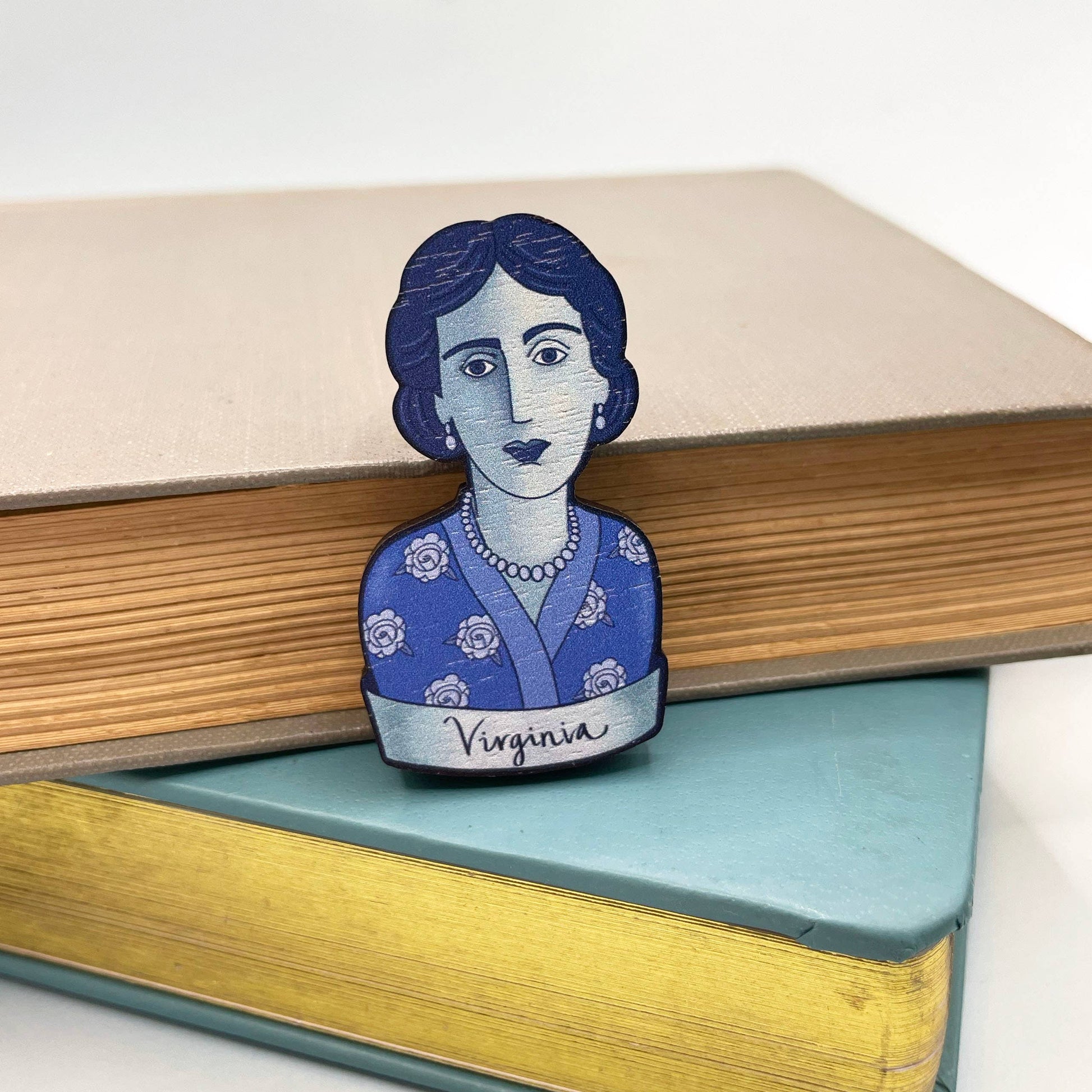 A wooden brooch depicting Virginia Woolf with a blue dress and rose motifs, secured by a metal bar pin sitting on a stack of books
