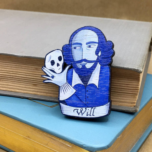 A wooden pin badge featuring William Shakespeare holding a skull, placed on an open book.