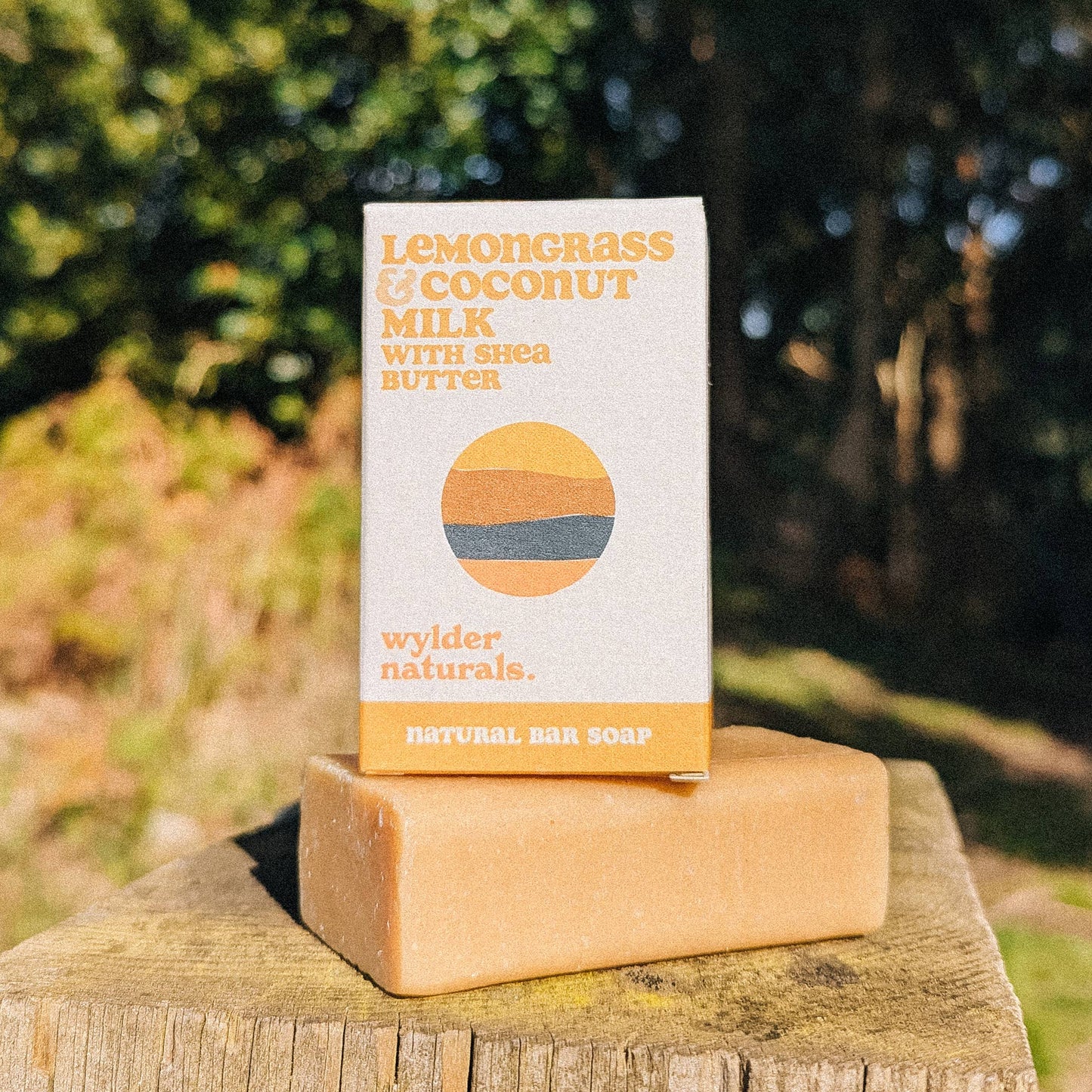 A bar of soap with packaging that reads 'Lemongrass & Coconut Milk with Shea Butter Natural Bar Soap'. The packaging is placed on a wooden surface with nature in the background.