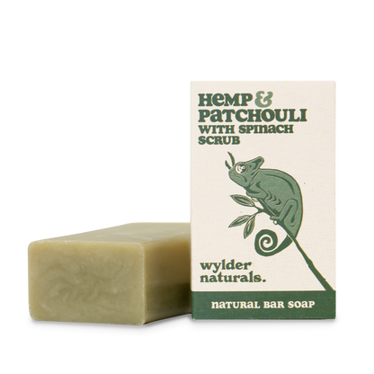 A bar of natural soap with a greenish hue, next to its packaging which features a green chameleon and text detailing the product as 'Hemp & Patchouli with Spinach Scrub'. The brand is 'Wylder Naturals' and the type as 'Natural Bar Soap'.